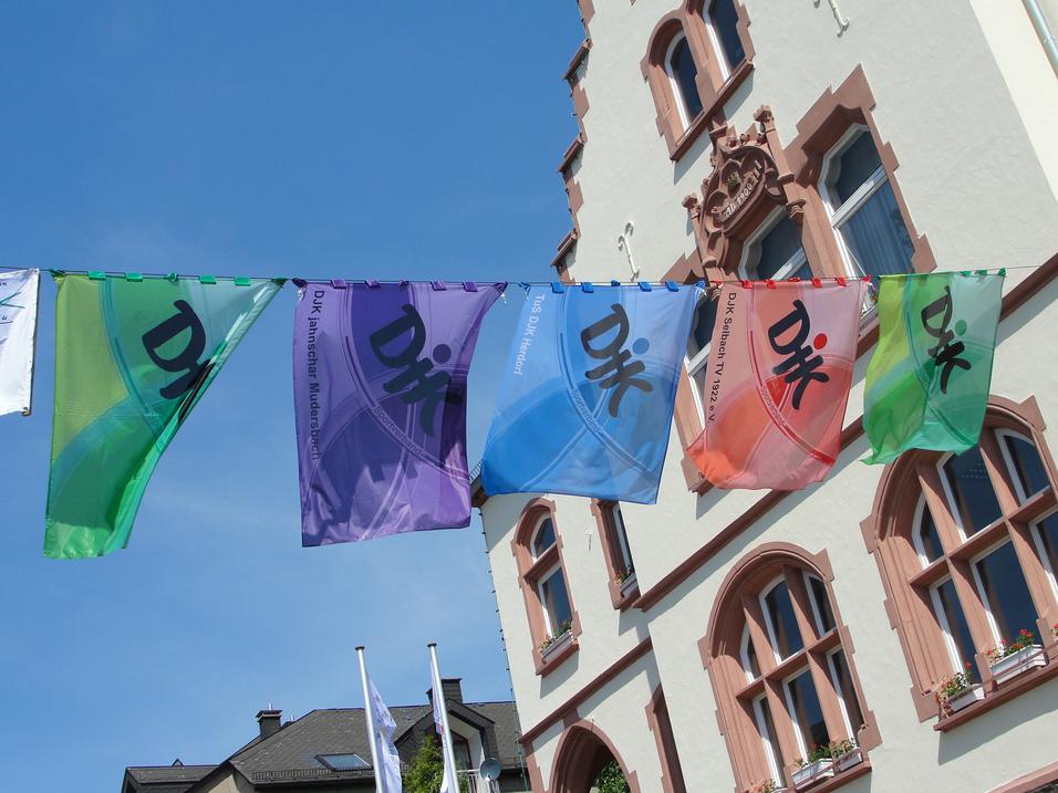 Farbenfrohe Fahnen mit Schriftzeichen h&auml;ngen zwischen historischen Geb&auml;uden in einem sonnigen Stadtbild.