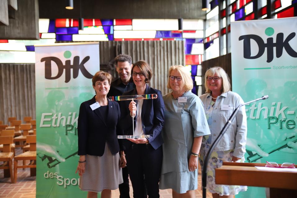 Gruppenfoto mit f&uuml;nf Personen vor einer farbigen Fenstergestaltung, die einen Preis in der Hand h&auml;lt, umgeben von DJK-Logos.