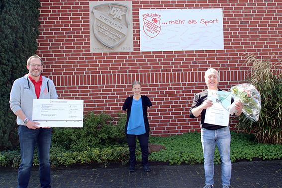 Zwei Personen halten Schecks und Urkunden vor einer Backsteinwand, w&auml;hrend eine dritte Person zwischen ihnen steht.