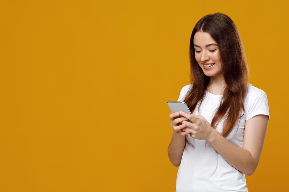 Junge Frau mit langen Haaren betrachtet l&auml;chelnd ihr Smartphone vor einem gelben Hintergrund.