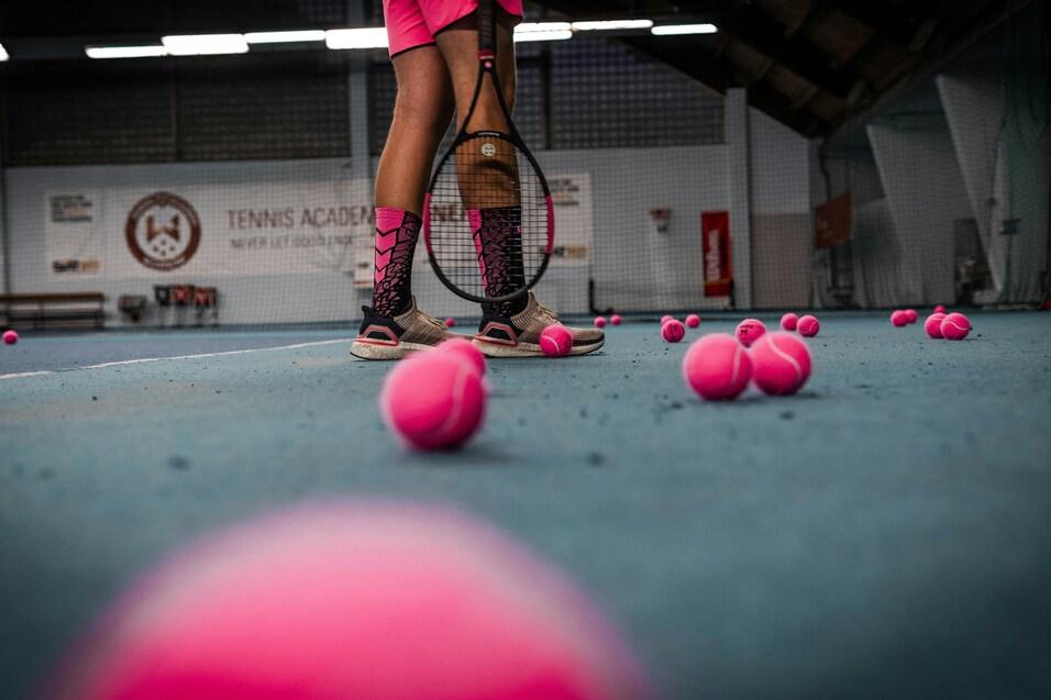 Nahaufnahme der Beine eines Tennisspielers in pinken Shorts und Socken, umgeben von vielen pinken Tennisb&auml;llen auf dem Boden.