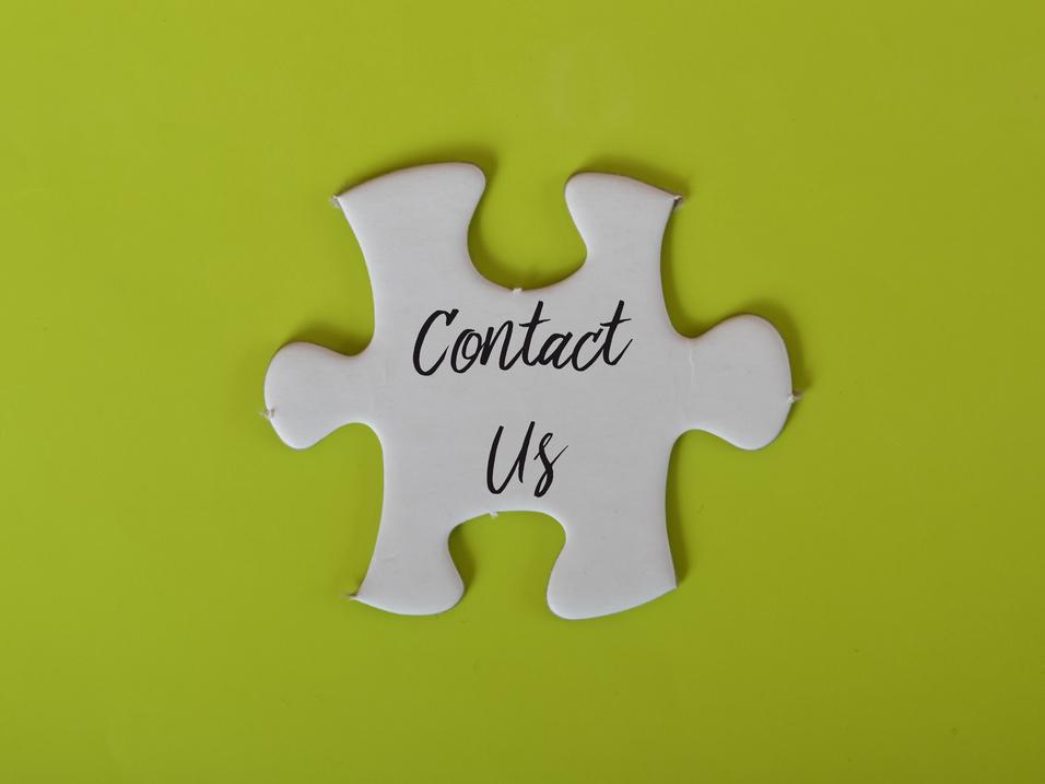 Wei&szlig;es Puzzlest&uuml;ck mit der Aufschrift &bdquo;Contact Us&ldquo; auf einer gr&uuml;nen Hintergrundfl&auml;che.