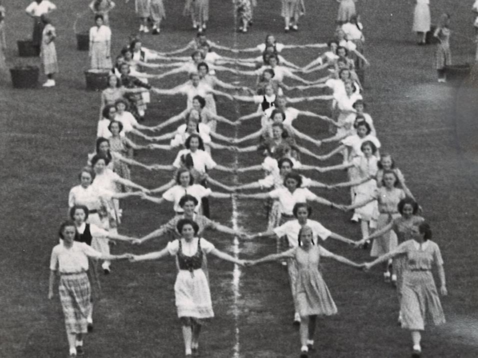 Gruppenauftritt von Frauen in historischen Kleidern auf einem Sportplatz, verbunden durch eine Kette aus H&auml;nden.