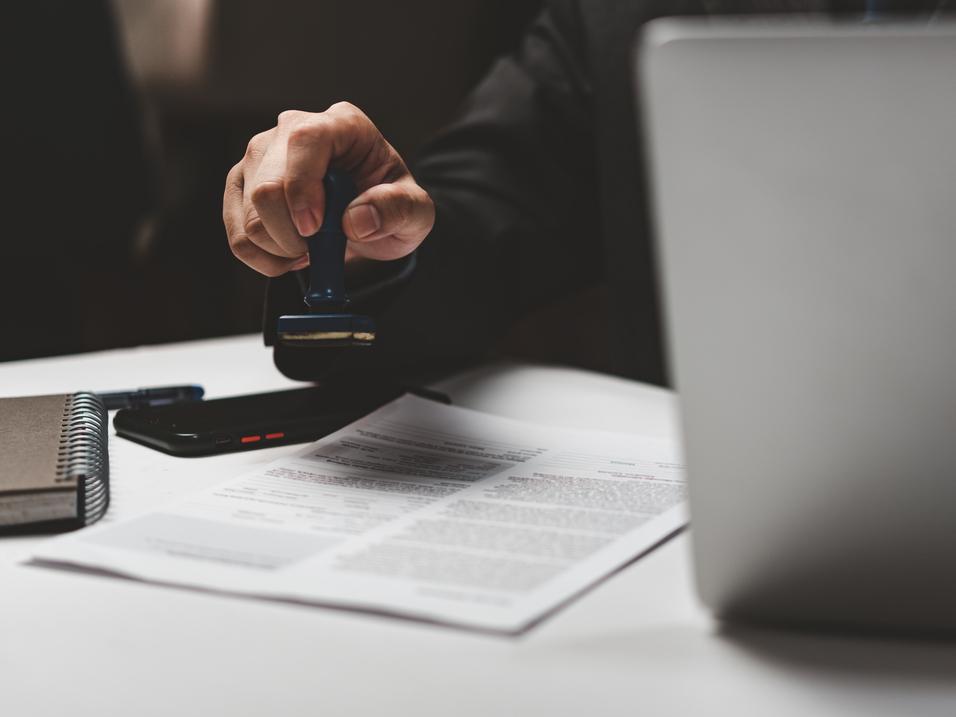 Hand mit Dokumentenstempel steht &uuml;ber einem Vertragsdokument und einem Laptop auf einem Tisch. Schwarz-wei&szlig;e Notizbuch liegt daneben.