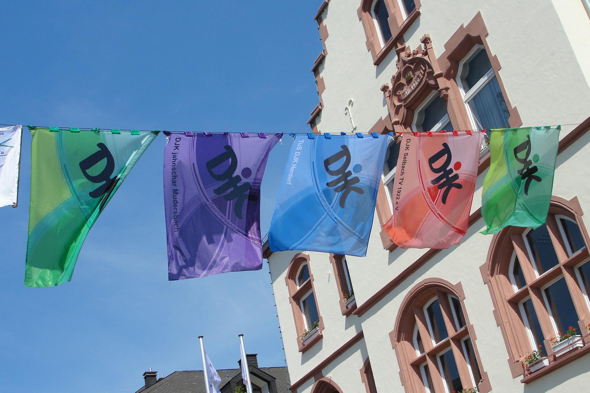 Farbenfrohe Fahnen mit Schriftzeichen h&auml;ngen zwischen historischen Geb&auml;uden in einem sonnigen Stadtbild.
