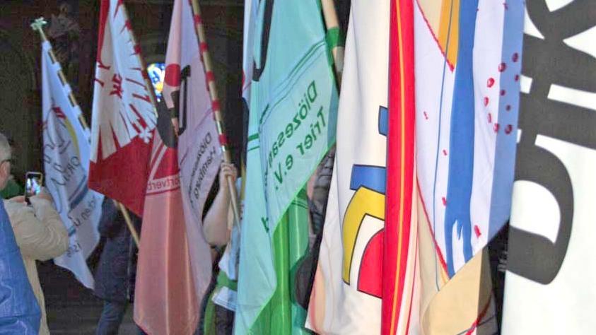 Fahnen unterschiedlicher Farben und Designs stehen in einem Kircheninneren, im Hintergrund ein gro&szlig;es Glasfenster.