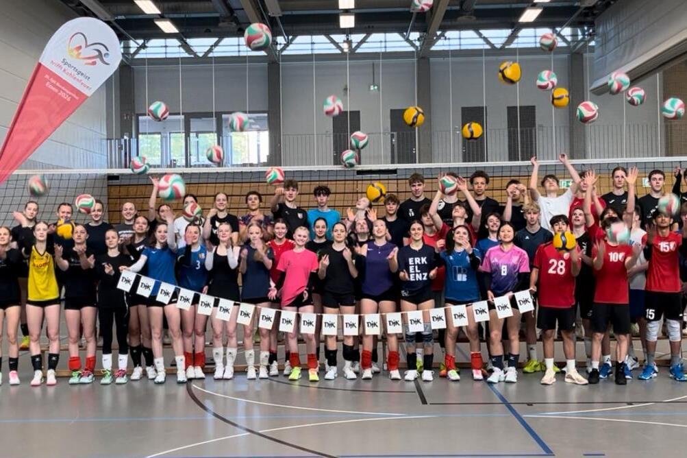 Gruppenshot von jungen Volleyballspielern, die jubelnd mit B&auml;llen in der Sporthalle stehen. Banner im Hintergrund.