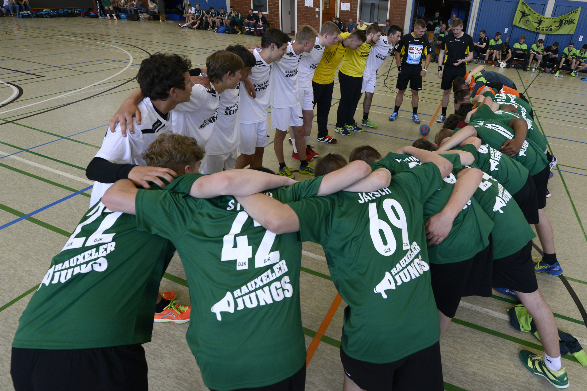 Zwei Gruppen von Jugendspielern in Sportanz&uuml;gen bilden einen Kreis in einer Sporthalle und bereiten sich auf ein Spiel vor.
