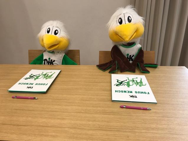 Zwei Stoffpuppen mit Vogelk&ouml;pfen sitzen an einem Holztisch, vor ihnen liegen Notizbl&ouml;cke und pinke Kugelschreiber.