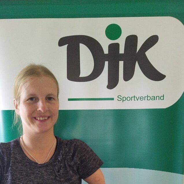 Portr&auml;t einer l&auml;chelnden Frau vor einem Banner mit dem Logo des DJK Sportverbands in gr&uuml;nen Farben.