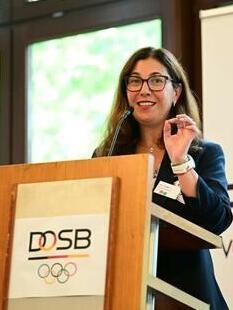 Frau spricht am Rednerpult bei der Frauen-Vollversammlung des DOSB, im Hintergrund das Banner mit dem Logo.