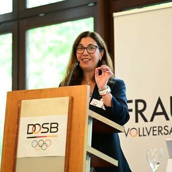 Frau spricht am Rednerpult bei der Frauen-Vollversammlung des DOSB, im Hintergrund das Banner mit dem Logo.