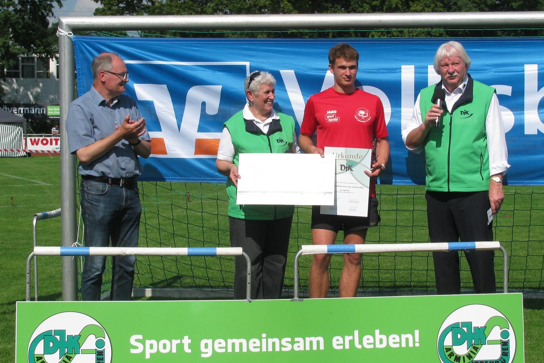 Vier Personen stehen am Fu&szlig;balltor und pr&auml;sentieren eine Auszeichnung auf einem Sportplatz, im Hintergrund ein Werbebanner.