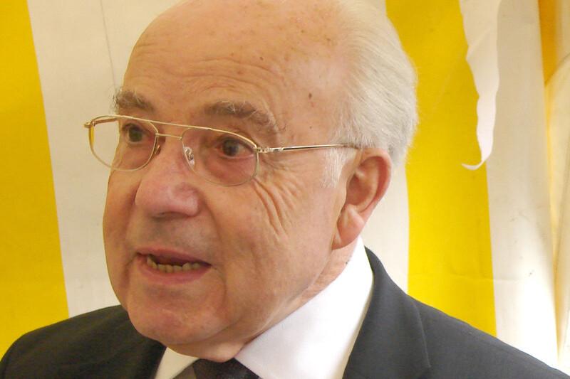 &Auml;lterer Mann mit Brille spricht vor einer gelben und wei&szlig;en gestreiften Hintergrundplane.