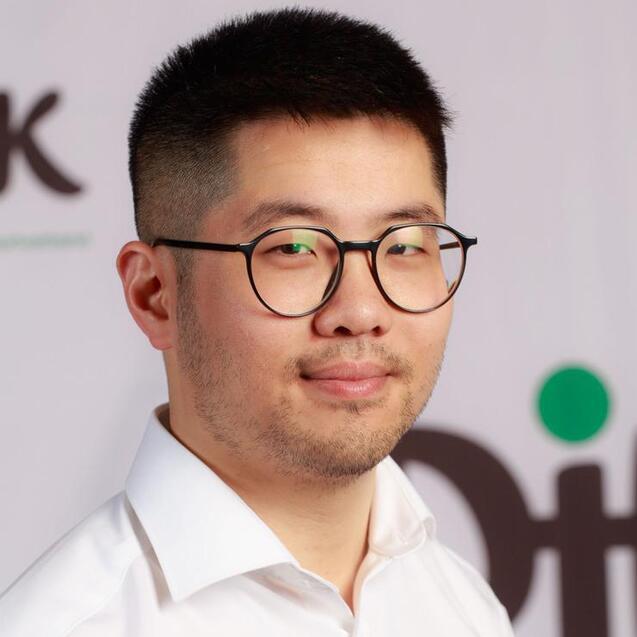 Porträt eines jungen Mannes mit Brille, in einem weißen Hemd mit einem Logo, vor einem unklaren Hintergrund.