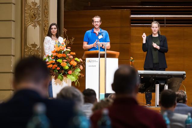 Drei Personen auf einer B&uuml;hne w&auml;hrend einer Veranstaltung, ein Mann spricht am Rednerpult, zwei Frauen stehen daneben.
