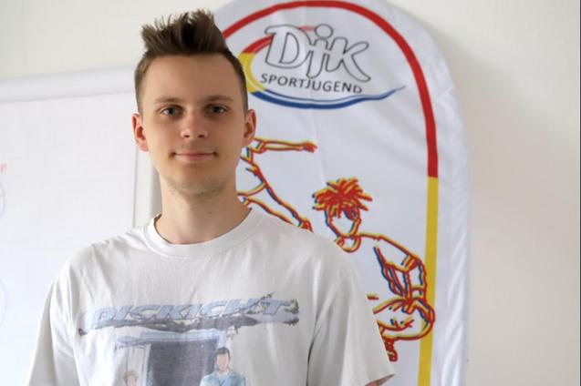 Junger Mann in T-Shirt steht vor einem Banner der DJK Sportjugend mit Sportmotiven.