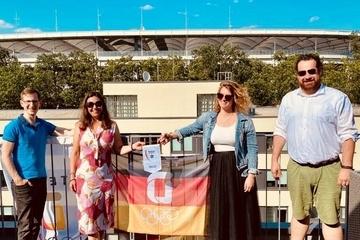 Vier Personen stehen auf einer Terrasse vor einer gro&szlig;en Flagge und zeigen ein Zertifikat, mit Stadion im Hintergrund.