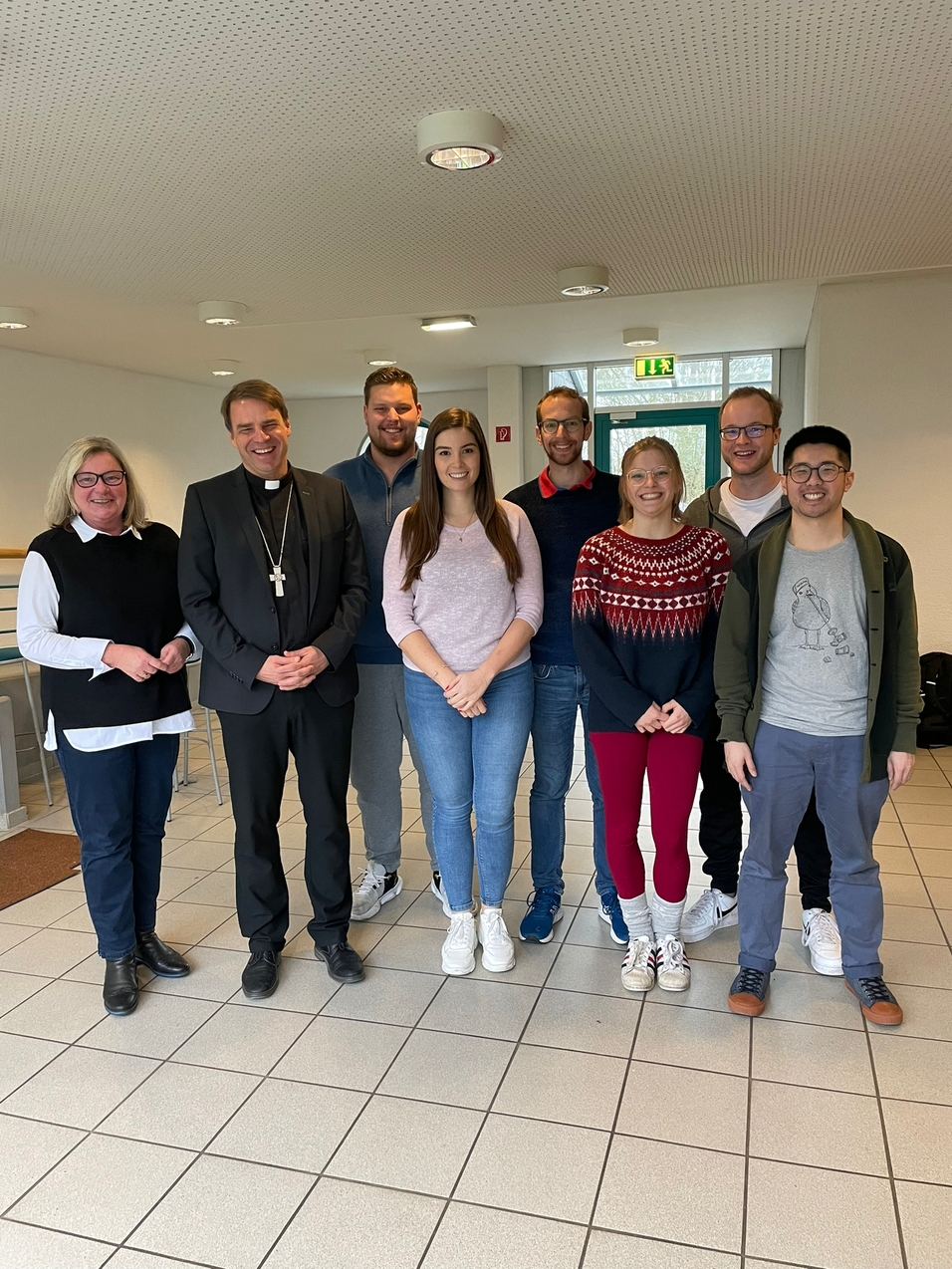 Gruppe von acht Personen, darunter ein Mann im Anzug mit Kleruskreuz, l&auml;cheln vor einem hellen Raum.