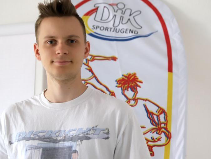 Junger Mann in T-Shirt steht vor einem Banner der DJK Sportjugend mit Sportmotiven.