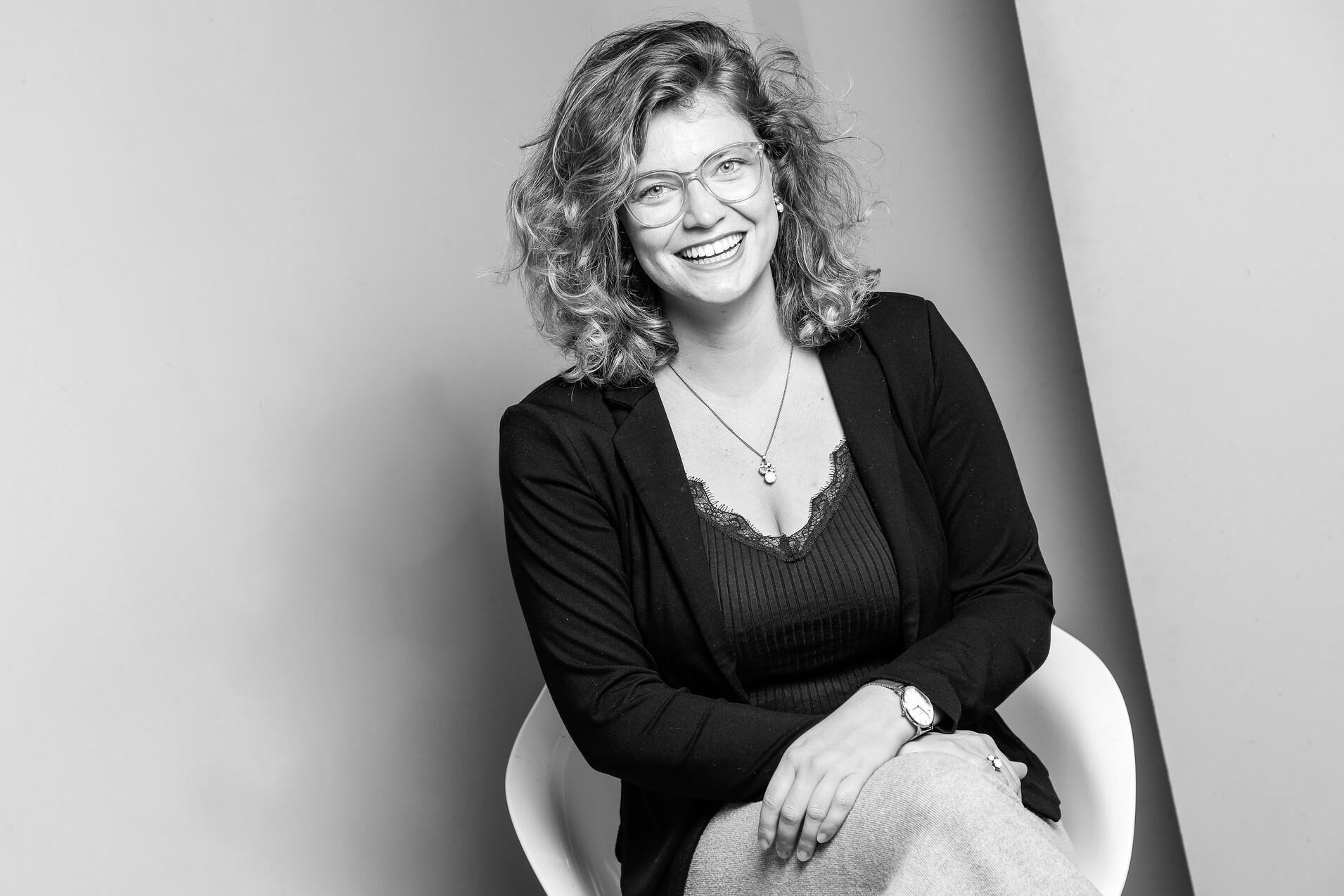 Portr&auml;t einer l&auml;chelnden Frau mit lockigem Haar, Brille und schwarzer Jacke, die auf einem wei&szlig;en Stuhl sitzt.