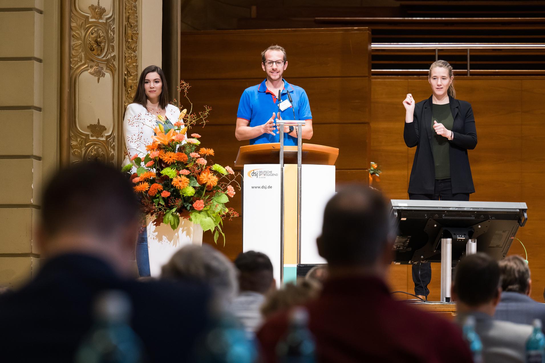 Drei Personen auf einer B&uuml;hne w&auml;hrend einer Veranstaltung, ein Mann spricht am Rednerpult, zwei Frauen stehen daneben.