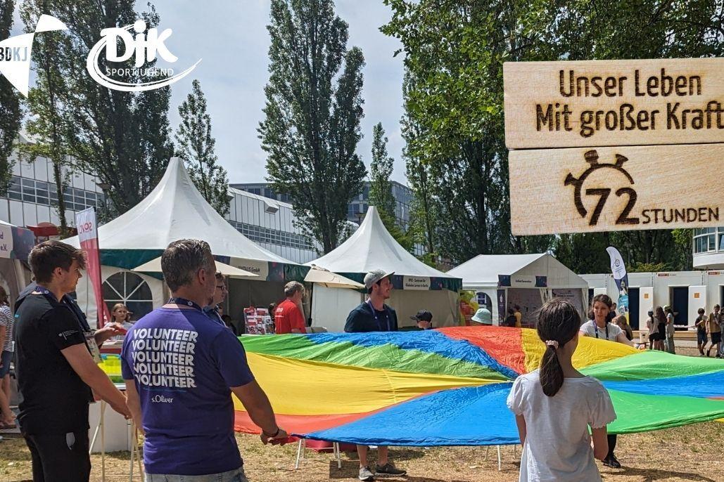 Gruppen von Menschen halten ein gro&szlig;es, buntes Fallschirmspielzeug auf einer Wiese bei einer Veranstaltung f&uuml;r Inklusion.