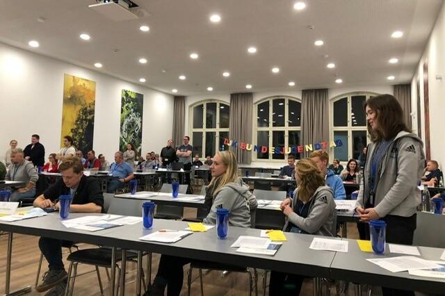 Gruppensitzung in einem hellen Raum mit mehreren Tischen, Teilnehmerinnen h&ouml;ren aufmerksam zu, Dekoration im Hintergrund.