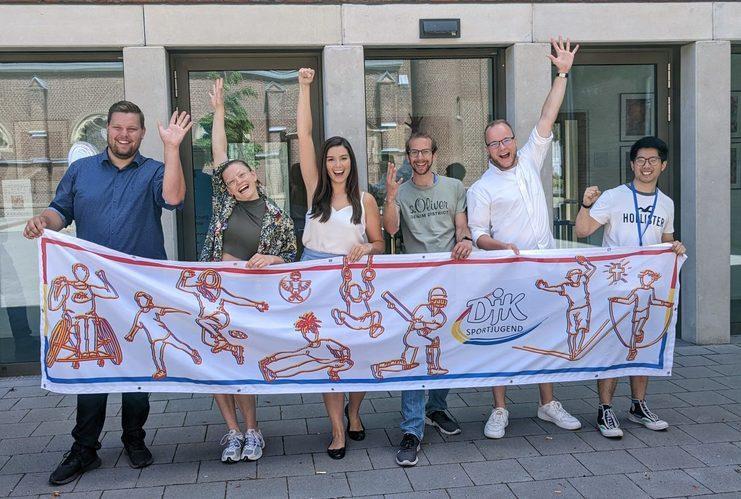 Gruppene foto von sechs Personen, die vor einem Geb&auml;ude ein Banner mit sportlichen Motiven halten und jubeln.