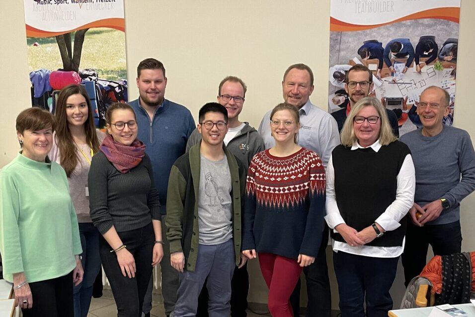 Gruppe von zw&ouml;lf Personen steht in einem Raum, l&auml;chelnd, hinter einem Tisch mit Namensschildern.