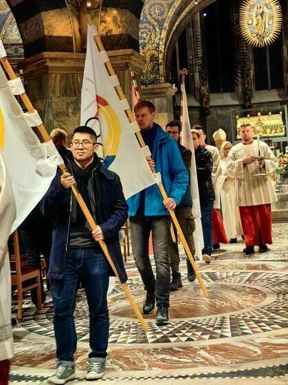 Menschen tragen Fahnen in einer Kirche w&auml;hrend eines feierlichen Anlasses, im Hintergrund eine Gruppe von Geistlichen.