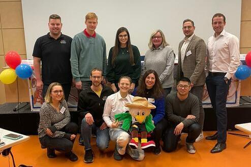 Gruppenevent mit zehn Personen und einem Maskottchen vor einer Leinwand, umgeben von bunten Luftballons.