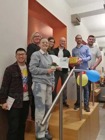 Gruppe stehender Personen auf einem Podest, einige halten Geschenke und Zertifikate in einer festlichen Umgebung.