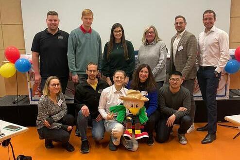 Gruppenevent mit zehn Personen und einem Maskottchen vor einer Leinwand, umgeben von bunten Luftballons.