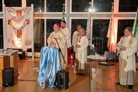 Ein Bischof steht an einem Pult, umgeben von weiteren Personen in liturgischen Gew&auml;ndern in einem festlichen Raum.