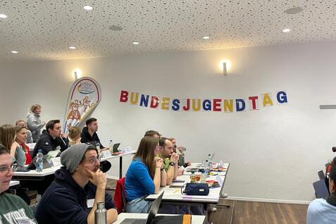 Teilnehmer eines Treffens sitzen an Tischen, w&auml;hrend ein Banner mit der Aufschrift &bdquo;BUNDESJUGENDTAG&ldquo; an der Wand h&auml;ngt.