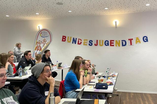 Teilnehmer eines Treffens sitzen an Tischen, w&auml;hrend ein Banner mit der Aufschrift &bdquo;BUNDESJUGENDTAG&ldquo; an der Wand h&auml;ngt.