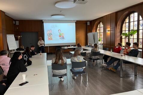 Gruppensitzung in einem hellen Raum mit Pr&auml;sentation und Teilnehmern, die einen Vortrag aufmerksam verfolgen.