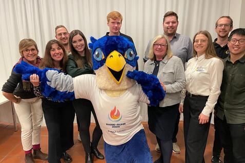 Gruppensfoto mit einem Maskottchen in blauer Federkleidung und zehn Personen, die l&auml;cheln und gemeinsam posieren.