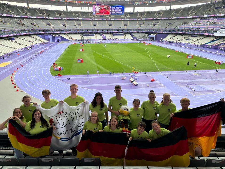 Gruppe junger Sportler in gr&uuml;nen T-Shirts h&auml;lt Deutschlandflaggen und ein Banner in einem Stadion mit Laufbahn.