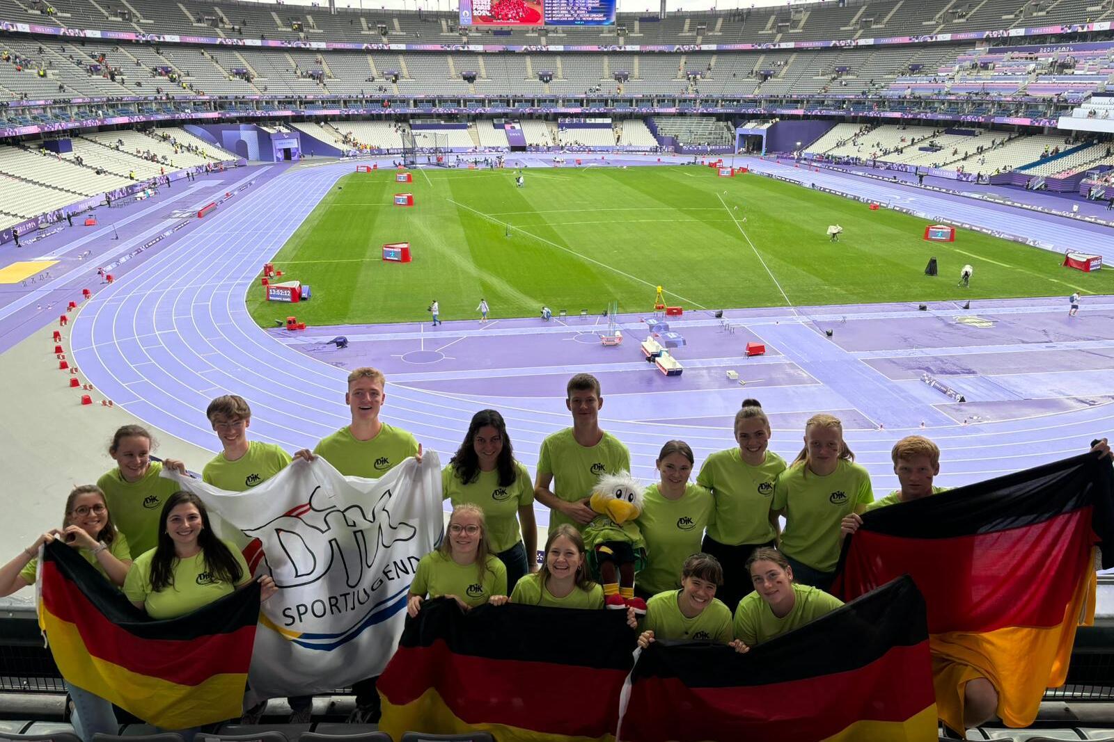 Gruppe junger Sportler in gr&uuml;nen T-Shirts h&auml;lt Deutschlandflaggen und ein Banner in einem Stadion mit Laufbahn.