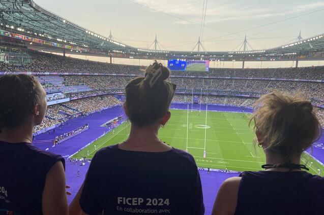 Drei Personen mit R&uuml;ckenansicht in lila Shirts beobachten ein Sportereignis in einem vollbesetzten Stadion.