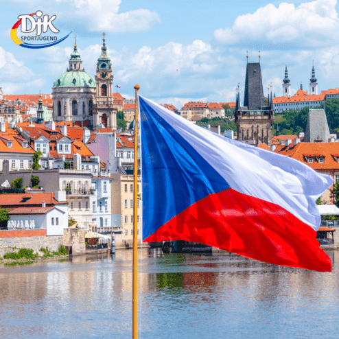 Flagge der Tschechischen Republik weht vor einer malerischen Stadtansicht mit historischen Geb&auml;uden und einem Fluss.