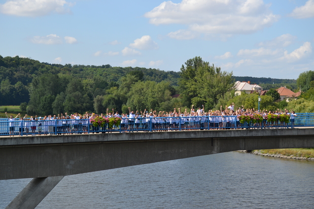 Vielz&auml;hlige Menschen stehen auf einer Br&uuml;cke und heben die Arme vor einer gr&uuml;nen Landschaft und einem Fluss.