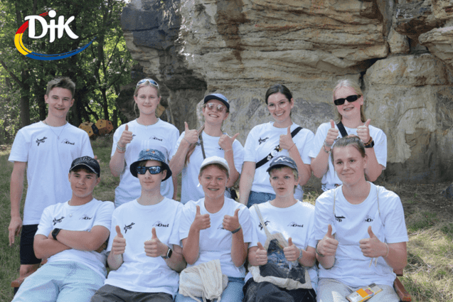 Gruppe von Jugendlichen in wei&szlig;en T-Shirts zeigt Daumen hoch, sitzend auf einer Bank vor Felsen im Freien.