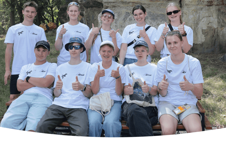 Gruppe von Jugendlichen in wei&szlig;en T-Shirts zeigt Daumen hoch, sitzend auf einer Bank vor Felsen im Freien.