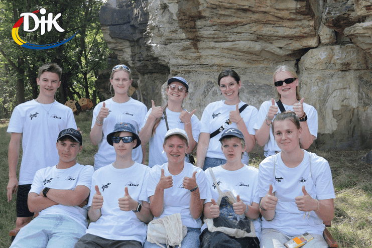 Gruppe von Jugendlichen in weißen T-Shirts zeigt Daumen hoch, sitzend auf einer Bank vor Felsen im Freien.