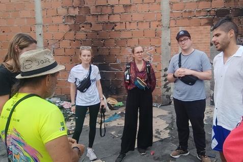 Gruppe von sechs Personen steht in einer urbanen Umgebung, vor einer rotbraunen Backsteinwand, bei informellem Austausch.