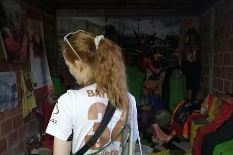 Junge Frau mit langen, lockigen Haaren tr&auml;gt ein Fu&szlig;balltrikot und schaut in einen chaotischen Raum voller Kleidung und Masken.