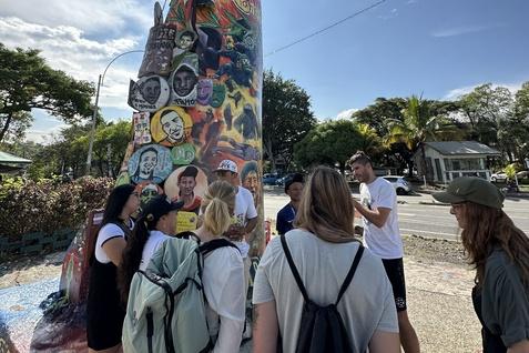 Gruppe von Menschen steht um eine bunte, graffitibespr&uuml;hte Statue mit verschiedenen Bildern und Aufschriften.